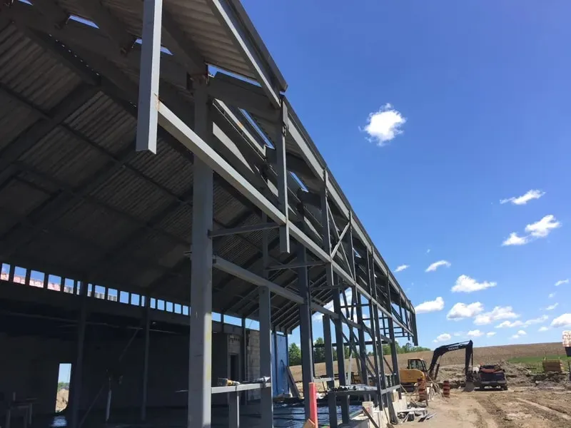 A steel frame going up on site: columns and beams with roof decking over the near bay, and an excavator working on the ground beyond it.