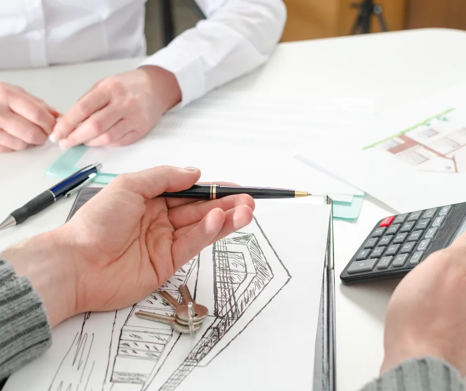 Two people at a table working over a hand-drawn building elevation, with a pen, a set of keys and a calculator beside the drawings.